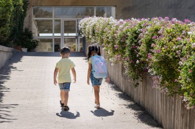 Okula geri dönen küçük bir çocuk. Kravat boyalı sırt çantalı küçük bir kız kardeşiyle okula yürür..