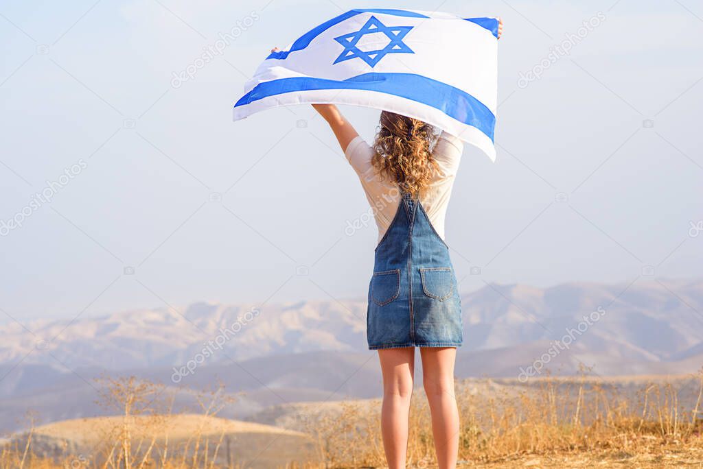 Teen curly hair Jewish girl standing with the flag of Israel on the amazing valley and blue sky background. Memorial day-Yom Hazikaron,Patriotic holiday Independence day Israel Yom Haatzmaut concept.