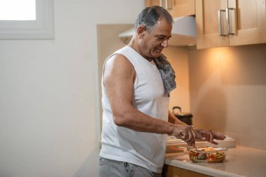 Active smiling athletic happy senior man standing in the kitchen and preparing salad.