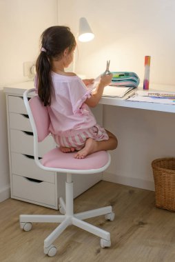 Uzun siyah elbiseli sevimli küçük kız modern odada elinde makasla beyaz masadaki pembe öğrenci sandalyesinde oturuyor..