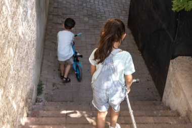 Bisikletli çocuk ve motosikletli kız kardeşi şehirde merdivenlerden aşağı iniyorlar..