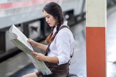 Bekleyen Asyalı hamile turist bankta oturmuş tren istasyonunda kırmızı bavullu haritaya bakıyor, yaz tatilinde sırt çantalı gezgin Tayland konsepti.