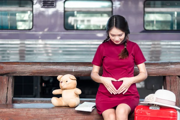 アジアの女性妊娠中の赤いドレステディベアとベンチに座ってハート型の手作りと鉄道駅の旅行で赤いスーツケースと地図 — ストック写真