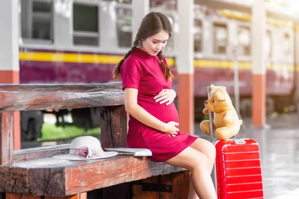 赤い荷物を運ぶベンチに座っているアジアの女性妊娠中の赤いドレスとテディベア鉄道駅の旅行で 夏の休暇のコンセプトでバックパックを持つ旅行者タイ — ストック写真