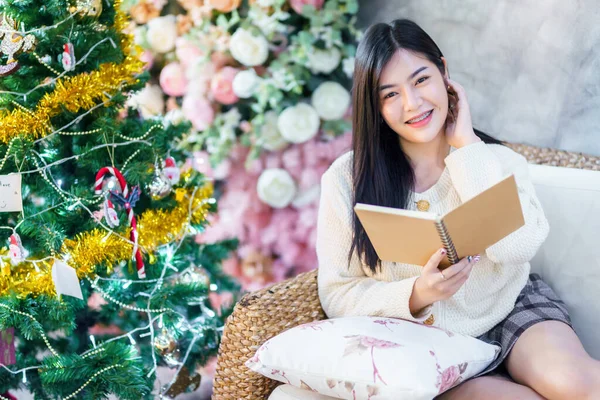 肖像画かわいい美しい肯定的な笑顔若いアジアの女性が自宅のリビングルームで本を読むの保持室内装飾クリスマスの間にX マスと新年の休日 — ストック写真