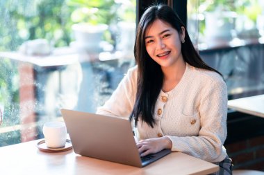 Serbest portre işi güzel pozitif gülümseme genç Asyalı kadın internette dizüstü bilgisayarla çalışıyor ve evde, oturma odasında ya da kafede kahve fincanı var, Business Lifestyle