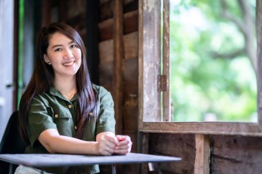 Mutlu portre mutlu mutlu gülümseyen güzel Asyalı kadın kahverengimsi yeşil elbise giyen şık hippi kahve fincanı olan ahşap bir evin arka camının yanında
