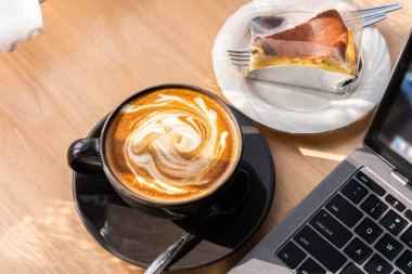 Sütlü sütlü sütlü sütlü sıcak kahve fincanı ve ev yapımı çikolatalı kek ve ahşap masanın üstünde dizüstü bilgisayar. İş konsepti sırasında bir kafede kahvaltı olarak.