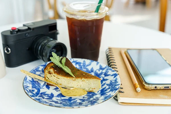 Americano iced black coffee in cup mug ,burnt face cheesecake placed and camera, smartphone in notebook on wood desk on top view. As breakfast In a coffee shop at the cafe,during business work concept