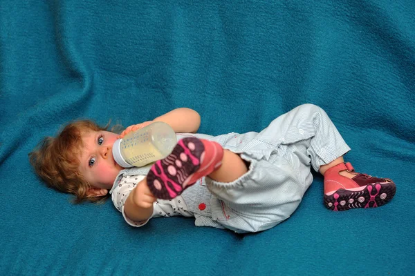 Kleines Mädchen liegt auf blauem Sofa und trinkt Milch aus Kolben — Stockfoto