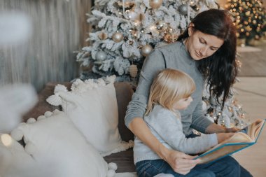 Anne ve kızı Noel ağacının yanında kitap okuyorlar.