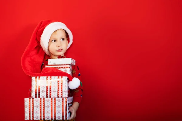 Gülen komik çocuk Noel Baba şapkası takıyor ve elinde kırmızı arka planda 2022 yılını kutlayan bir sürü hediye kutusu tutuyor. Mutlu Noel hediyeleri alışverişi.