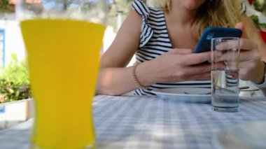 Çizgili elbiseli tanınmaz sarışın kadın ya da telefonda tişört saati, SMS okuyor, eğlence içeriğini izliyor. Ön planda bir bardak portakallı soda var. Cep telefonuyla kahvaltı..