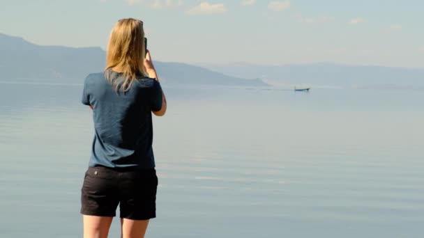 Donna bionda irriconoscibile in camicia blu e pantaloncini corti neri usando il suo telefono per scattare foto di lago o mare. barca a remi naviga in lontananza e si possono vedere le montagne. Lady scatta foto su smartphone. — Video Stock