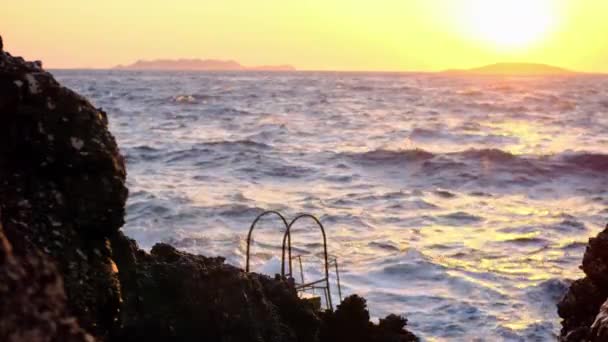 Cinematic widoki szalejącego falistego oceanu lub morza w falach o zachodzie słońca, z gór na pierwszym planie i drabiny zejść do wody do pływania. Fale rozbijają się o skały na tle świtu. — Wideo stockowe