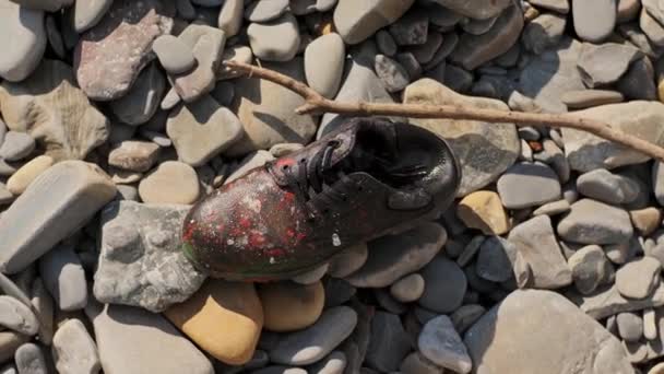 Stary skórzany but, pokryty farbą, leży na kamiennej plaży. Odpady wyrzucane i zanieczyszczające środowisko. Ekologia potrzebuje pomocy. pojęcie lekceważenia zachowania życia na planecie. — Wideo stockowe