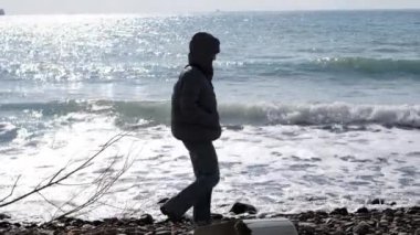 Ceketli, kotlu, deniz ya da okyanus boyunca taş plaj boyunca yürüyen tanınmayacak kadar yalnız bir kadın. Dışarısı soğuk ve rüzgarlı, bayan başlık ve kalın bir ceket giyiyor. Yalnız düşünme konsepti.