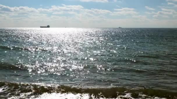 Prachtig uitzicht op zee of oceaan met kleine golven, zon reflecteert off-oppervlakte van het water. Op de achtergrond zijn schepen en olietankers te zien in de verte, die op weg is naar de haven. Mooie wolkenlucht. — Stockvideo