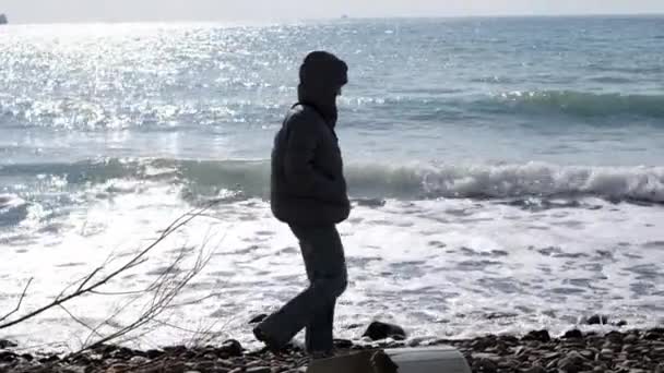 Oigenkännlig ensam kvinna i dunjacka och jeans promenader längs havet eller havet längs sten stranden. Det är kallt och blåsigt ute, damen bär huva och varm jacka. begreppet tänkande ensam. — Stockvideo