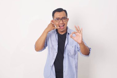 İzole edilmiş beyaz arka planda durup gülümseyen Asyalı genç adam el ve parmaklarıyla telefon hareketi yapıyor.