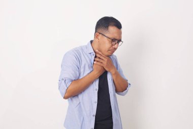 Boğazı ağrıyan, boynuna dokunan, beyaz arka planda sıradan kıyafetler giyen genç bir adam.