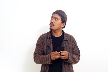 Geleneksel kostüm giymiş Asyalı bir adam elinde cep telefonuyla karışık bir ifadeyle bekliyor.