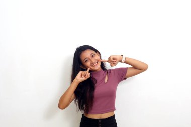 Gülümseyen genç bir kadın, beyaz arka planda iki parmağıyla yanağını gösteriyor.