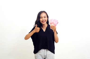 Asyalı genç bir kadın elinde Endonezya parası tutarken ve baş parmağını kaldırırken ayakta duruyor. İzole edilmiş