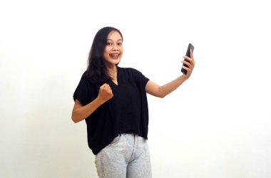Joyful asian young woman standing while holding a cellular phone. Good news concept.