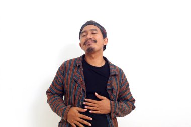 Asyalı adam karnına dokunurken ayakta duruyor, tok ve tatmin olmuş hissediyor. Beyaz arkaplanda izole