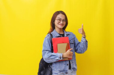 Elinde kitap tutarken gülümseyen Asyalı genç bir kadın baş parmak işareti yapıyor. İzole edilmiş