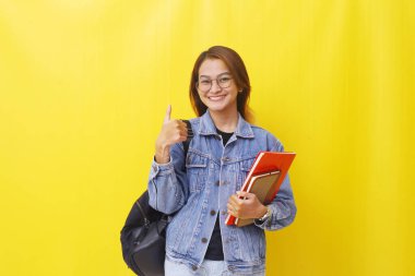 Asyalı genç bir üniversite öğrencisi elinde kitaplarla ayakta duruyor ve başparmağını kaldırıyor. Sarıda izole edilmiş.