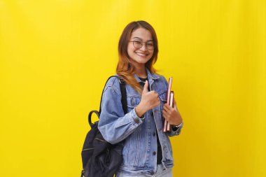 Asyalı genç bir üniversite öğrencisi elinde kitaplarla ayakta duruyor ve başparmağını kaldırıyor. Sarıda izole edilmiş.