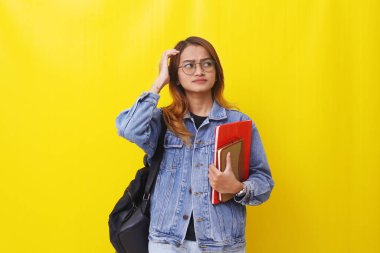 Düşünceli Asyalı genç bayan kitap ve okul çantasıyla ayakta duruyor. Sarıda izole edilmiş..
