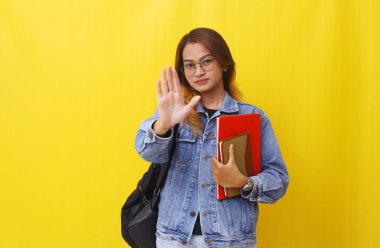 Asyalı genç bir üniversite öğrencisi elinde kitaplarla ayakta dikiliyor. El hareketlerini durduruyor. Sarıya sarılmış.