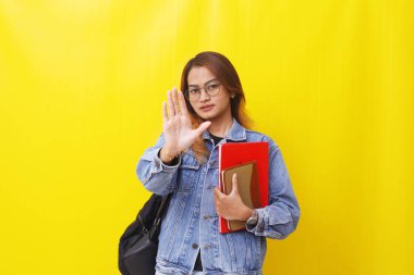 Asyalı genç bir üniversite öğrencisi elinde kitaplarla ayakta dikiliyor. El hareketlerini durduruyor. Sarıya sarılmış.