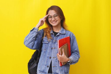 Asyalı genç bir üniversite öğrencisi elinde kitaplarla ayakta durup el hareketleriyle selam veriyor.