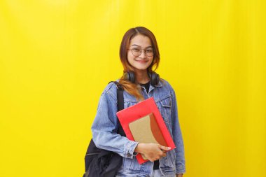 Asya 'nın genç ve güzel üniversitesi elinde kitap ve okul çantasıyla ayakta duruyor. Sarıda izole edilmiş.