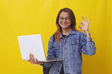 Mutlu Asyalı genç üniversite öğrencisi elinde dizüstü bilgisayarla el hareketleriyle ayakta duruyor.