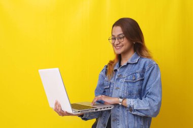 Elinde dizüstü bilgisayarla duran mutlu Asyalı genç bir üniversite öğrencisi. Sarı arkaplanda izole edilmiş