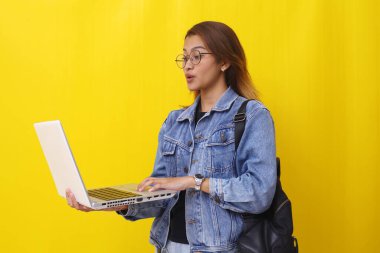 Elinde dizüstü bilgisayarla dikilen Asyalı genç bir üniversite öğrencisi. Sarı arkaplanda izole edilmiş