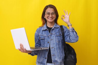 Neşeli Asyalı üniversite öğrencisi elinde dizüstü bilgisayarla el işaretleri ile duruyor.