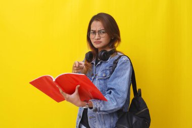 Asyalı hoşnutsuz üniversite öğrencisi bir kitabı işaret ederken ayakta duruyor. Sarı arkaplanda izole edilmiş