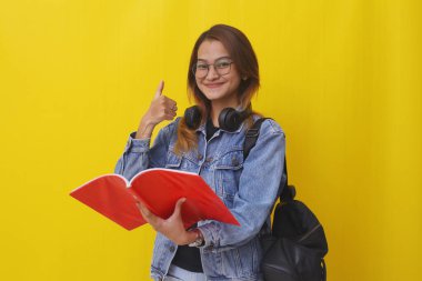 Asyalı üniversite öğrencisinin baş parmağını kaldırırken elinde bir kitap tutmasının portresi. Sarıda izole edilmiş.