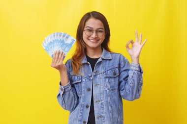 Elinde Endonezya parası olan gülümseyen Asyalı genç bir kadın el hareketi yapıyor.