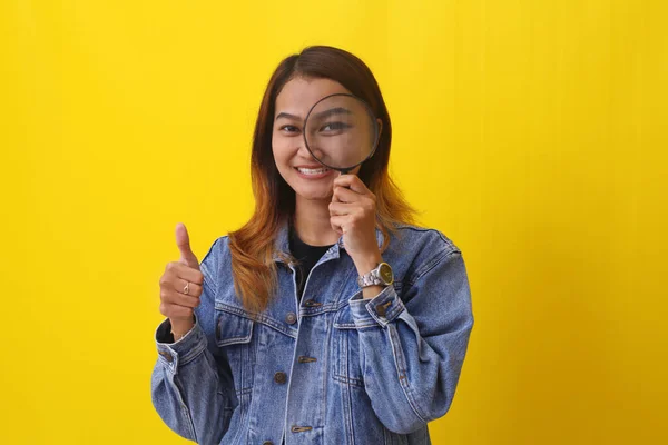 年轻的亚洲女人举着放大镜 同时竖起大拇指 在黄色背景下隔离 — 图库照片