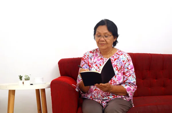 Ηλικιωμένη Ασιάτισσα Που Κάθεται Στον Καναπέ Ενώ Γράφει Στο Βιβλίο — Φωτογραφία Αρχείου