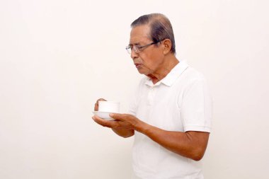 Elderly asian man holding a cup of coffee or tea while blows it. Isolated on white background