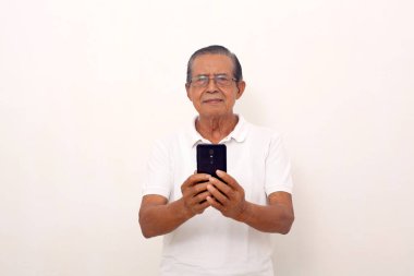 Calm elderly asian man standing while holding a cell phone. Isolated on white background