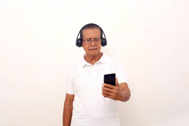 Asian old man standing while listening music from his phone using headphone. Isolated on white